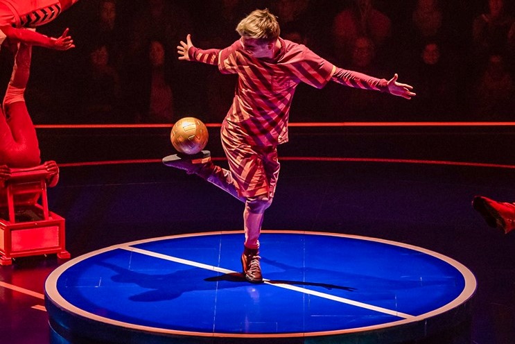 Team Fayolle Un artiste de cirque en costume rayé jongle avec un ballon sur un disque lumineux.