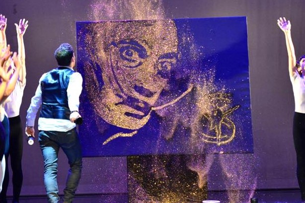 Théo Normand Un artiste crée un portrait en poussière colorée sur scène, entouré de danseurs.