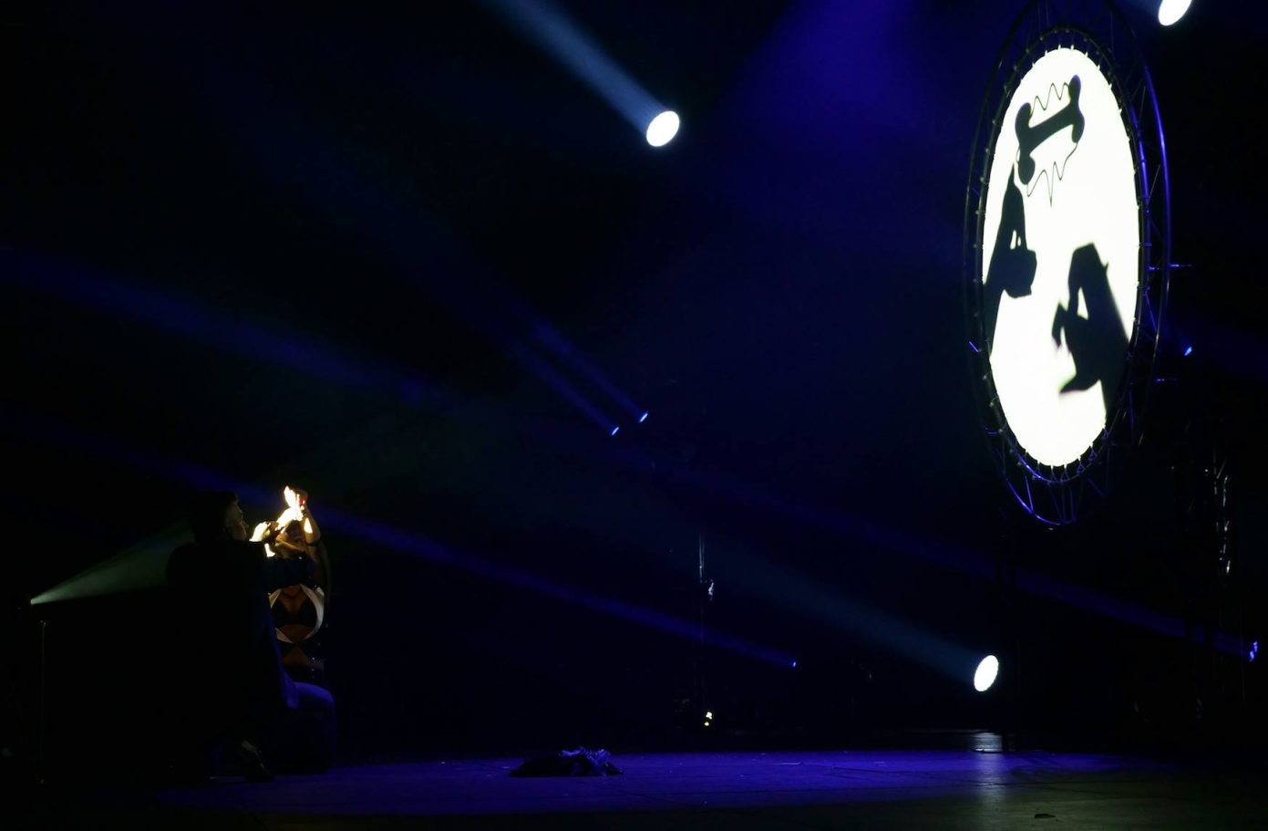 Black fingers Artiste sur scène éclairé par des lumières, avec un grand projecteur en arrière-plan.