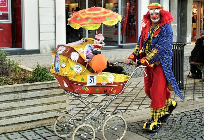 Clown en déambulation Clown coloré poussant une poussette décorée de plusieurs accessoires et jouets.