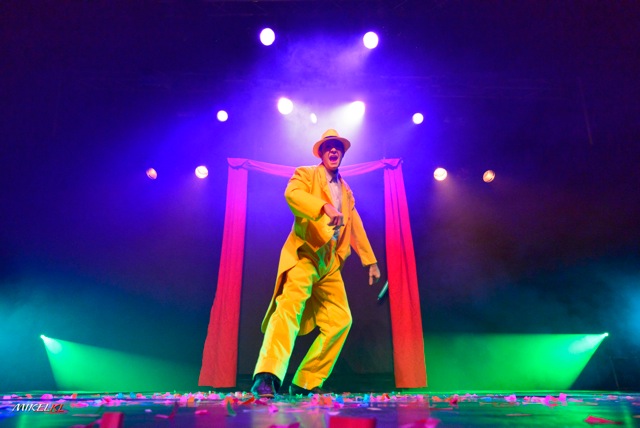 Maxime Minerbe Artiste en costume jaune sur scène, illuminé par des lumières colorées.